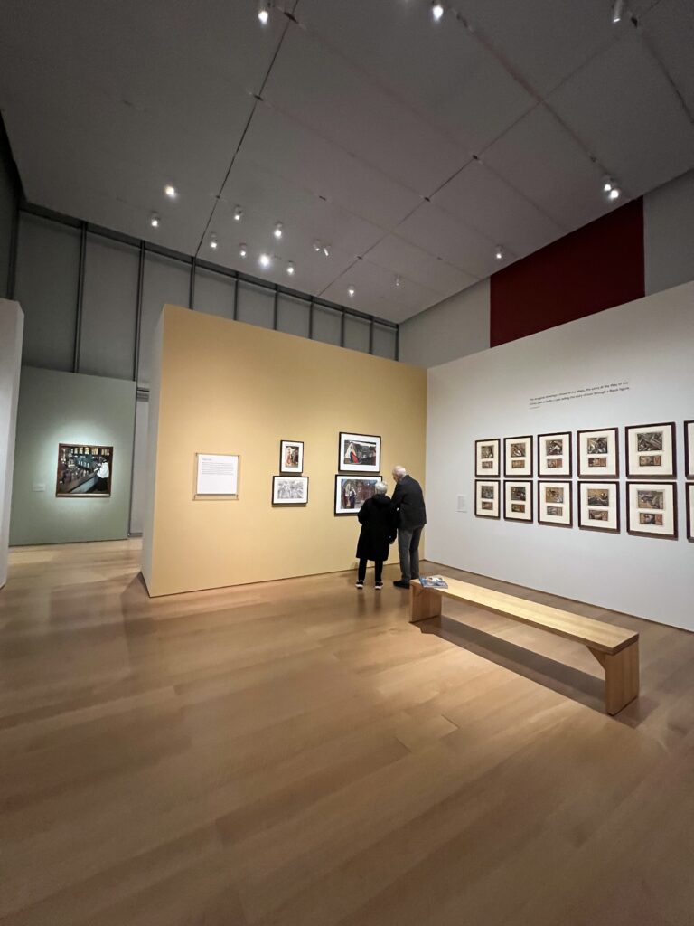 Gallery installation displaying four religious oil paintings on a yellow wall adjacent to a 12 panel liturgical work on a white wall with strong overhead lighting