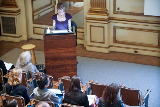A photograph of a woman presenting to a seated audience