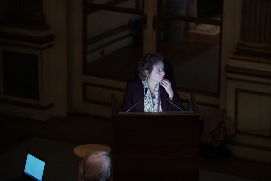 A photograph of a woman presenting in a dark room