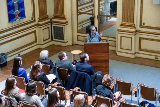 A photograph of a woman delivering a presentation to a seated audience