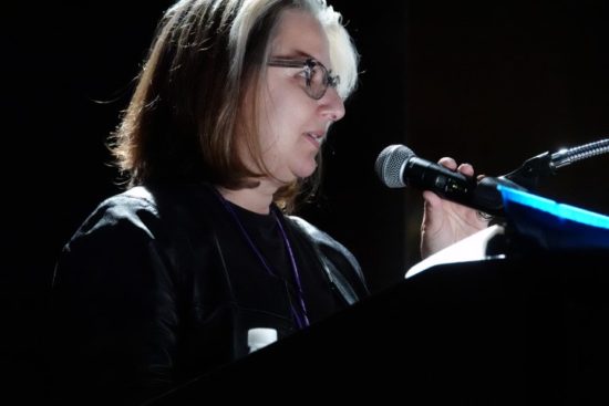 A photograph of Lena Stringari behind a microphone in a dark space