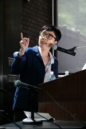 A photograph of Hiroko Kimura-Myokam standing behind a podium looking up