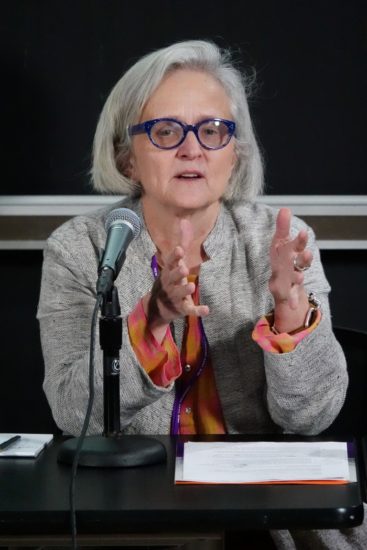 A photograph of Jill Sterrett sitting at a table with a microphone at the NYU "It's About Time!" Symposium