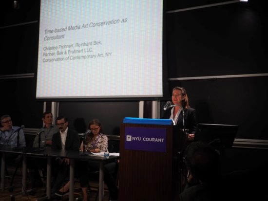 A photograph of Christine Frohnert giving a presentation with a projection screen at the NYU "It's About Time!" Symposium