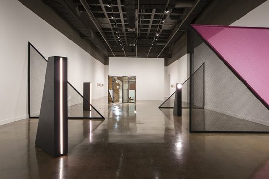 A photograph of a white walled gallery with large black and pink geometric sculptures