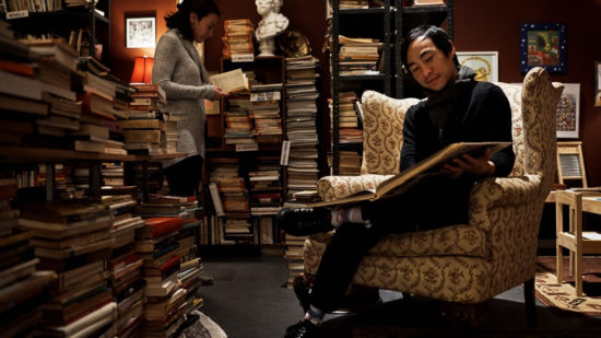 A man sits in a bookstore where the shelves are covered floor to ceiling with books, reading an oversize book.