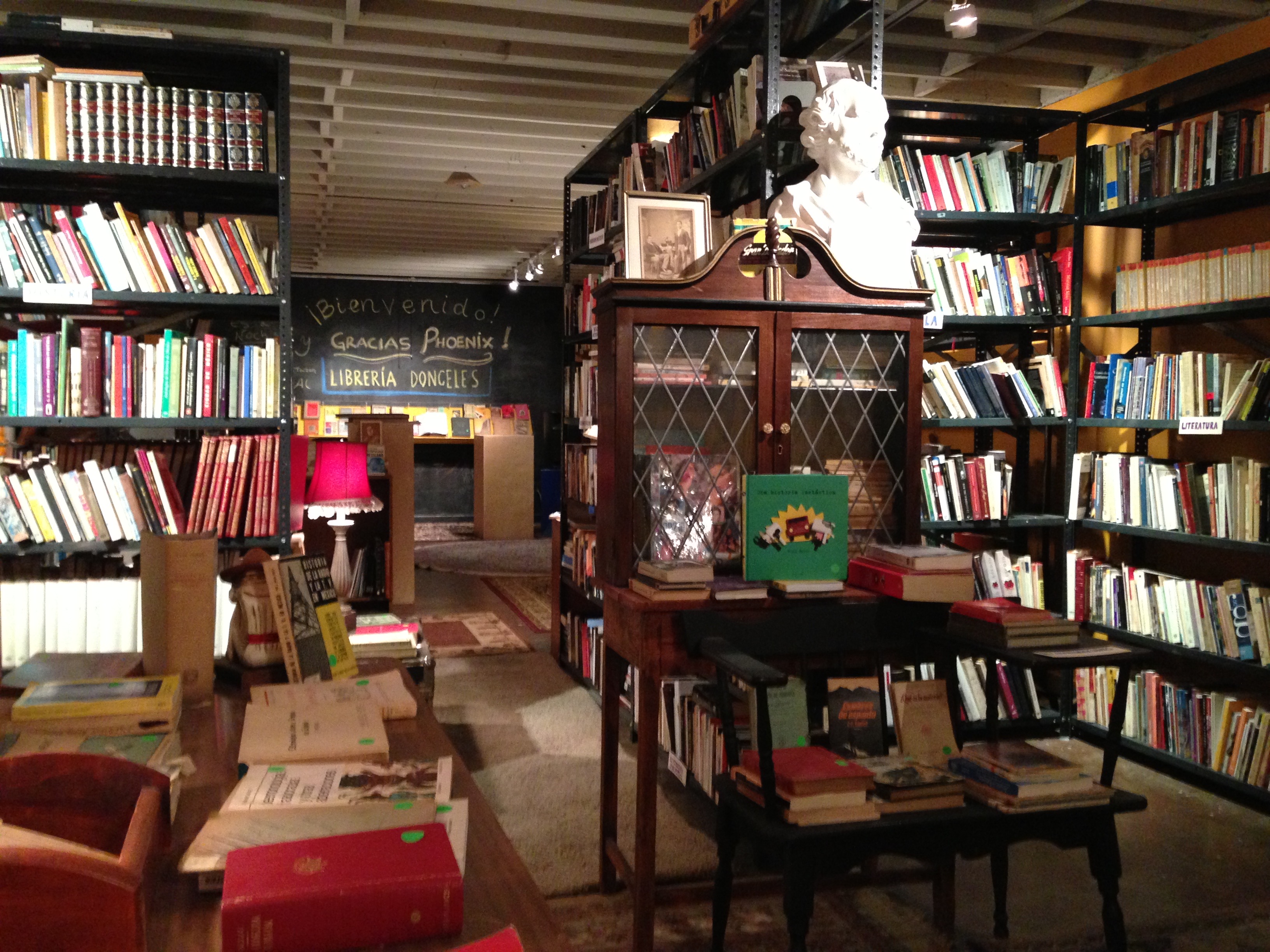 A room filled with bookshelves and bookcases leading to a second room displaying a chalkboard displaying “Bienvenido! Gracias Pheonix! Libreria Donceles”.