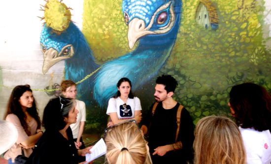 A group of women surround Maria Chatzidakis interviewing Heitor Correa in front of his mural of two peacocks.