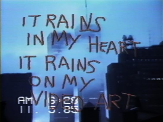 A film still showing the World Trade Towers, time stamped AM 6:28, 11.5.85, overlaid with the words "IT RAINS IN MY HEART IT RAINS ON MY VIDEO ART"