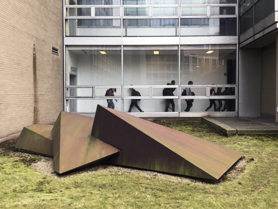 Students are visible walking through the hallway behind Beverly Pepper’s Trinity, 1971, which sits on the lawn between MIT Buildings 16 and 26.