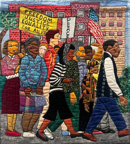 Quilt featuring diverse group marching at a protest holding an American flag and signs that read “Freedom Justice Equality for All Pe[ople]” and “Peace”