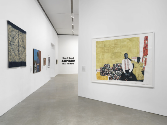 View of AAMARP exhibition with various artworks on white walls, including a textile piece, paintings, and a large framed artwork of two figures.