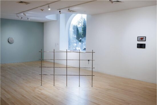 A gallery room contains a low metal grid structure positioned on a wooden floor, with a large arched window letting in natural light. The walls are mostly empty, with only a few small artworks visible.