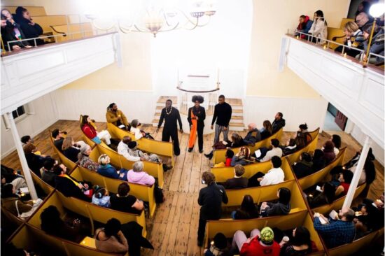 Photo from above showing a crowd watching three performers who are standing at the front of the room.