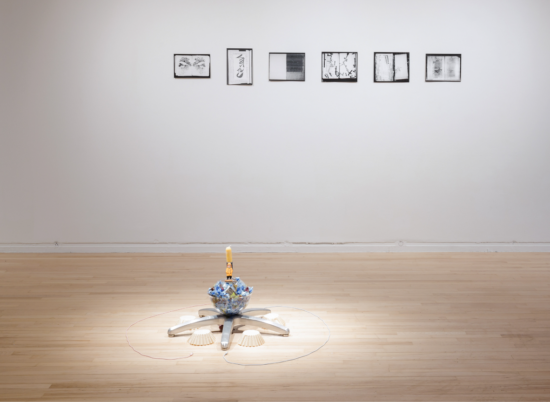 A small sculptural installation sits on the floor under a spotlight, surrounded by a circular arrangement of objects and wires. Behind it, a row of framed black-and-white drawings is mounted on the wall